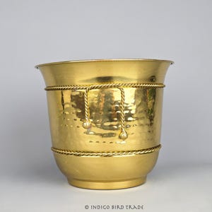 May include: A gold-coloured metal planter with a hammered texture. The planter has a flared top and a rounded base. Decorative rope trim and hanging accents add detail to the design. Suitable for indoor or outdoor use.
