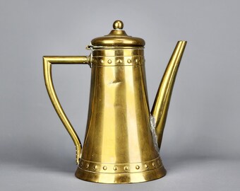Op de afbeelding: Een vintage messing koffiepot met een lange tuit en een handvat. De pot heeft een deksel met een kleine knop en is versierd met klinknagels aan de boven- en onderkant. De pot is gemaakt van glanzend messing en staat tegen een grijze achtergrond.