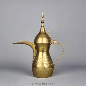 May include: A gold-colored, traditional Arabic coffee pot, or dallah, with a long spout and a curved handle. The pot features floral engravings and a tall, pointed lid. The background is a solid gray.