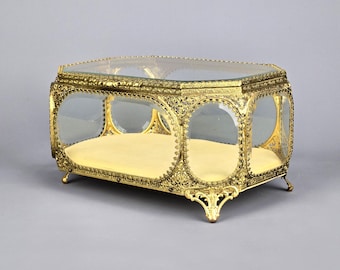 Könnte beinhalten: Eine goldfarbene Glas-Schmuckschatulle mit dekorativem Design und Samt-Auskleidung. Die Schatulle hat einen Klappdeckel und steht auf vier verzierten Füßen.