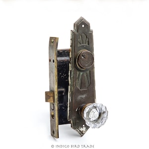 May include: An antique door lock set with a decorative, aged bronze finish. The set includes a rectangular backplate, a crystal doorknob, and a keyhole. The lock shows signs of wear, adding to its vintage appeal.