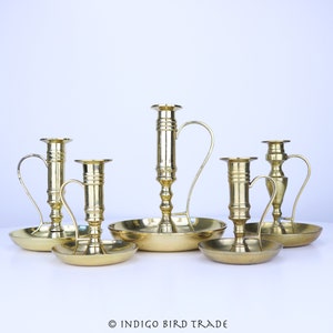 May include: Set of five brass candle holders in a variety of styles and sizes. The candle holders have a polished gold finish and are arranged on a white surface.