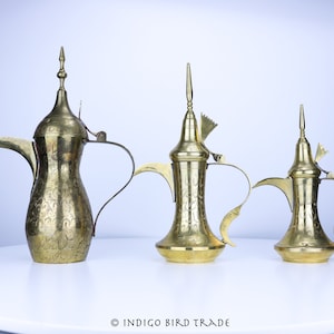 May include: Three gold-colored metal coffee pots with ornate designs and handles. The pots are arranged in a row on a white surface.  © INDIGO BIRD TRADE