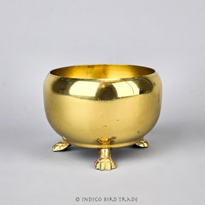 May include: A gold-colored metal bowl with a rounded shape and three small feet. The bowl is made of brass and has a shiny finish.