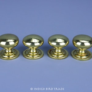 Könnte beinhalten: Vier goldfarbene Schrankknöpfe mit abgerundeter Oberseite und kreisförmiger Basis. Die Knöpfe haben eine glänzende, reflektierende Oberfläche und sind vor einem hellblauen Hintergrund platziert.