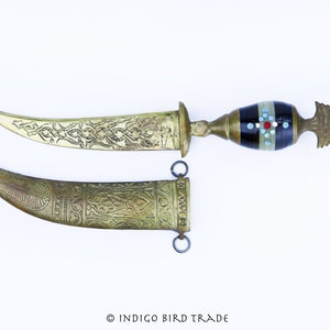 May include: A brass dagger with an ornate handle and a sheath. The handle is decorated with black and white enamel and red and blue accents. The blade is curved and has an intricate design.