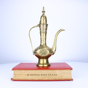 May include: A gold-colored metal teapot with intricate floral designs sits on top of a red book with the text "INDIGO BIRD TRADE" printed on the front.