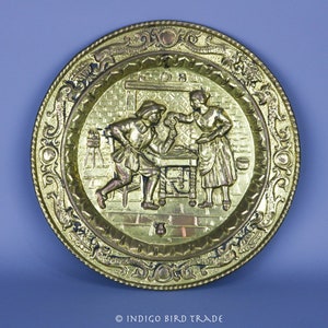 May include: A round, gold-colored metal wall plate with an embossed design of two figures in a scene. The plate has a decorative border with intricate details.