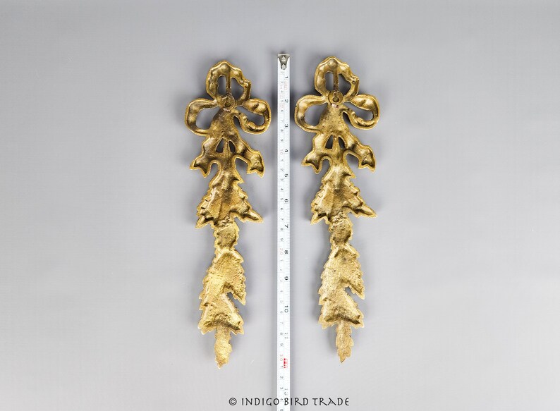 May include: Two gold-toned metal decorative pieces with a floral design. Each piece features a bow at the top and a series of cascading leaves.