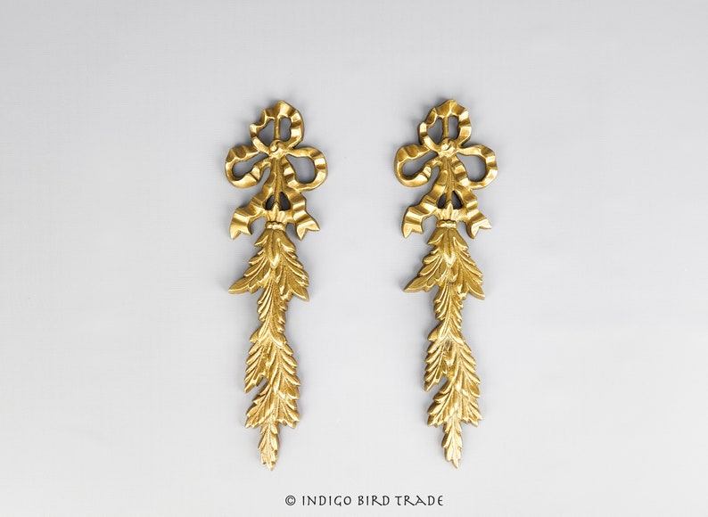 May include: A pair of gold-colored metal decorative ornaments with a bow and a cascading design. The ornaments are on a light gray background.