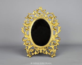 May include: An ornate, oval-shaped mirror with a decorative, scrolled frame in a gold tone. The mirror is on a stand and has a black reflective surface.