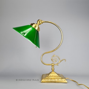 May include: A vintage desk lamp with a vibrant green glass shade and a brass-colored, curved arm. The lamp has a square base and a decorative, swirled design. The cord is visible, and the lamp is set against a neutral background.