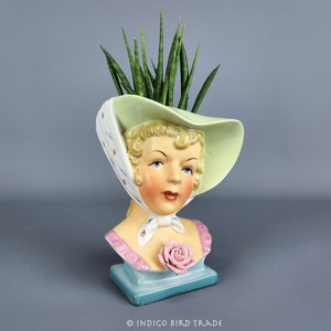 Op de afbeelding: Een vintage keramische plantenbak in de vorm van een vrouwenhoofd en -schouders, met een hoed en sjaal. De plantenbak is beschilderd met een lichtgroene hoed, een roze roos en een blauwe basis. De plantenbak bevat een groene plant.