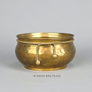 May include: A hammered brass bowl with a rope design around the rim and hanging tassels. The bowl has a rounded shape and a golden hue. The text "© INDIGO BIRD TRADE" is at the bottom.