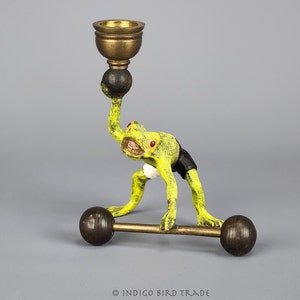 May include: A green frog figurine lifting a barbell with a gold candle holder on its head. The frog is wearing black shorts and a white band around its waist.