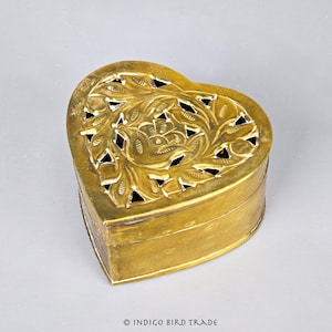 May include: A heart-shaped, brass-colored box with intricate floral cutouts on the lid. The box has a hinged design and a slightly aged appearance. The floral design is centered on the lid and is surrounded by geometric cutouts.