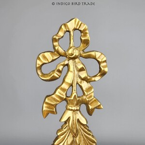May include: A gold-colored metal ornament shaped like a bow with a decorative, ornate design. The bow is tied with a ribbon and has a pointed tip.