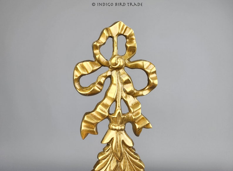 May include: A gold-colored metal ornament shaped like a bow with a decorative, ornate design. The bow is tied with a ribbon and has a pointed tip.