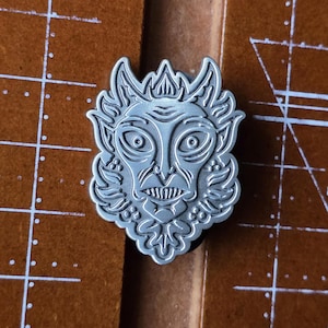 May include: A silver-colored metal pin featuring a detailed, stylized face with large eyes, a grimacing mouth, and ornate details. The pin has a textured surface and is likely a decorative accessory.
