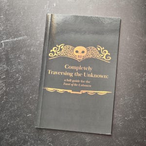 May include: A black book with a gold skull and wings design on the cover. The title is "Completely Traversing the Unknown: a full guide for the Tarot of the Unknown."
