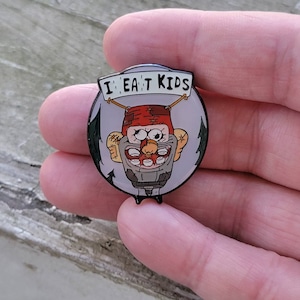 May include: A round enamel pin featuring a cartoon character with a red hat and a wide, toothy grin. The pin has the text "I EAT KIDS" on a banner above the character's head. The design is set against a light gray background with dark green accents.