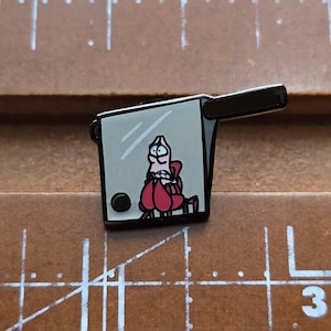 May include: A black and white enamel pin featuring a cartoon character inside a retro television set. The character is wearing a red outfit and appears to be distressed. The pin has a black handle and a black dot on the side.