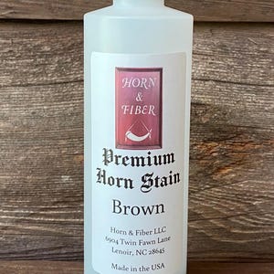 May include: A white plastic bottle with a black cap, labeled "Premium Horn Stain Brown." The label features the "Horn & Fiber" logo and company information. The bottle contains a brown liquid, and is made in the USA.