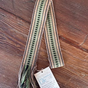 May include: A woven textile strip with a repeating pattern of black and green rectangles on a tan background. The fringe is green and tan. A tag reads "Early American Life magazine DIRECTORY OF TRADITIONAL AMERICAN CRAFTS 2025."