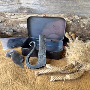 May include: A survival kit featuring a metal striker, flint pieces, and a small tin. The striker has a curved handle and a rectangular body with an engraved letter. A bundle of rope is also included.