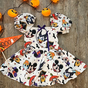 May include: A white dress with a Halloween theme featuring Mickey Mouse, Donald Duck, and Goofy. The dress has a purple ribbon at the neck and puff sleeves. The dress is covered in a pattern of the characters dressed in Halloween costumes.