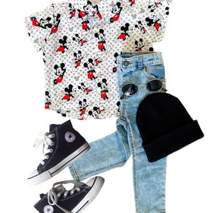 May include: A children's outfit featuring a white button-down shirt with a Mickey Mouse print, light blue jeans, black high-top sneakers, a black beanie, and sunglasses. The shirt has a classic collar and short sleeves. The sneakers have white laces and soles.