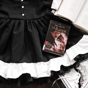 May include: A black dress with a white ruffle trim and a black lace overlay. A book titled "The Raven and Other Writings" by Edgar Allan Poe is lying on the dress. A pair of white sneakers are on the floor next to the dress.