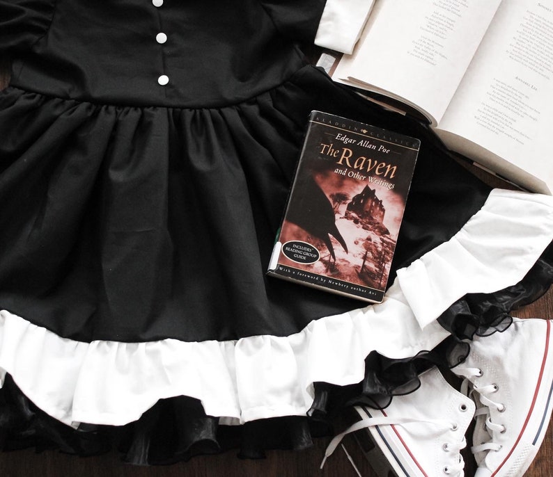 May include: A black dress with a white ruffle trim and a black lace overlay. A book titled "The Raven and Other Writings" by Edgar Allan Poe is lying on the dress. A pair of white sneakers are on the floor next to the dress.