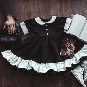 May include: A black dress with a white collar and ruffle trim, a black backpack, a book titled "The Raven and Other Poems" by Edgar Allan Poe, and white sneakers on a wooden floor.
