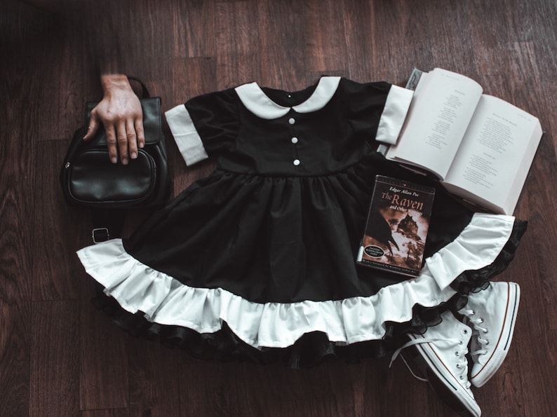 May include: A black dress with a white collar and ruffle trim, a black backpack, a book titled "The Raven and Other Poems" by Edgar Allan Poe, and white sneakers on a wooden floor.