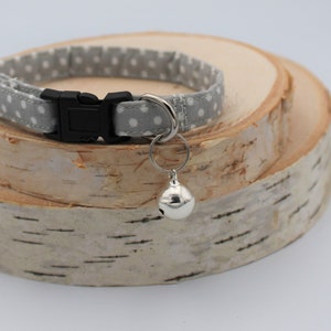 May include: A gray and white polka dot cat collar with a silver bell attached. The collar has a black plastic buckle.