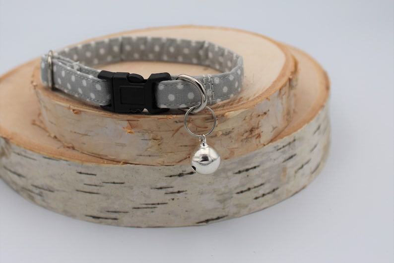 May include: A gray and white polka dot cat collar with a silver bell attached. The collar has a black plastic buckle.