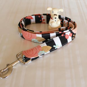 May include: A black and white striped dog leash with a floral pattern in shades of pink, gold, and green. The leash has a silver clip and is coiled up on a light brown surface.