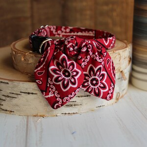 May include: Red bandana dog collar with a black buckle and a white paisley pattern. The collar is tied in a bow.
