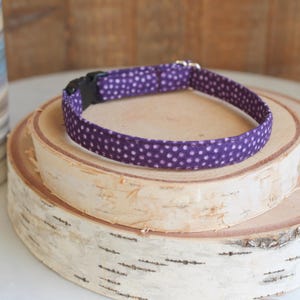 May include: A purple dog collar with white polka dots, featuring a black plastic buckle and silver-colored metal hardware. The collar is displayed on a stack of birch wood slices, with a decorative vase in the background.