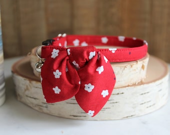 Op de afbeelding: Rode kattenhalsband met een wit bloemenpatroon en een bijpassende strik. De halsband heeft een bel.