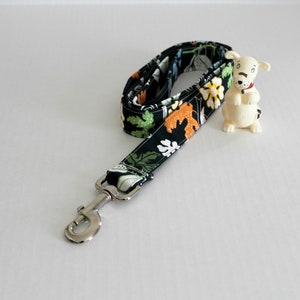 Op de afbeelding: Een zwarte hondenriem met een bloemenprint met groene, oranje en witte bloemen. De riem heeft een zilveren clip.