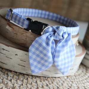 Puede incluir: Un collar de perro de gingham azul y blanco con una pajarita a juego. El collar tiene una hebilla de pl&aacute;stico negra.
