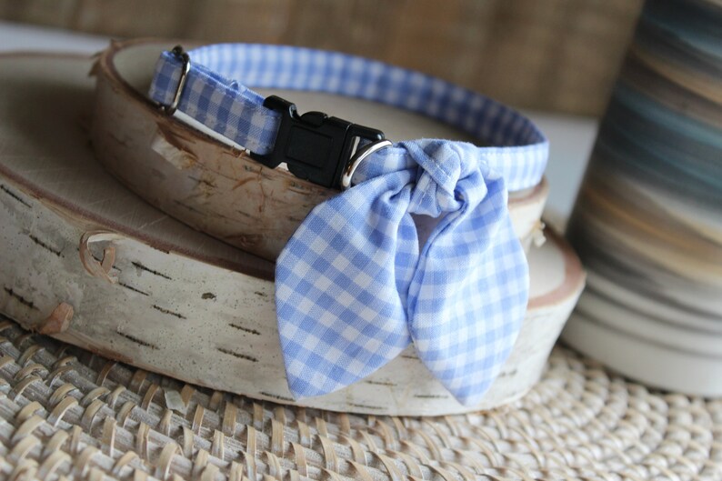Puede incluir: Un collar de perro de gingham azul y blanco con una pajarita a juego. El collar tiene una hebilla de pl&aacute;stico negra.