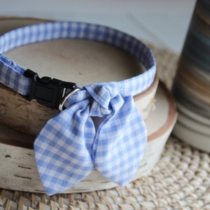 Puede incluir: Un collar de perro de gingham azul y blanco con una pajarita. El collar tiene una hebilla de pl&aacute;stico negra.
