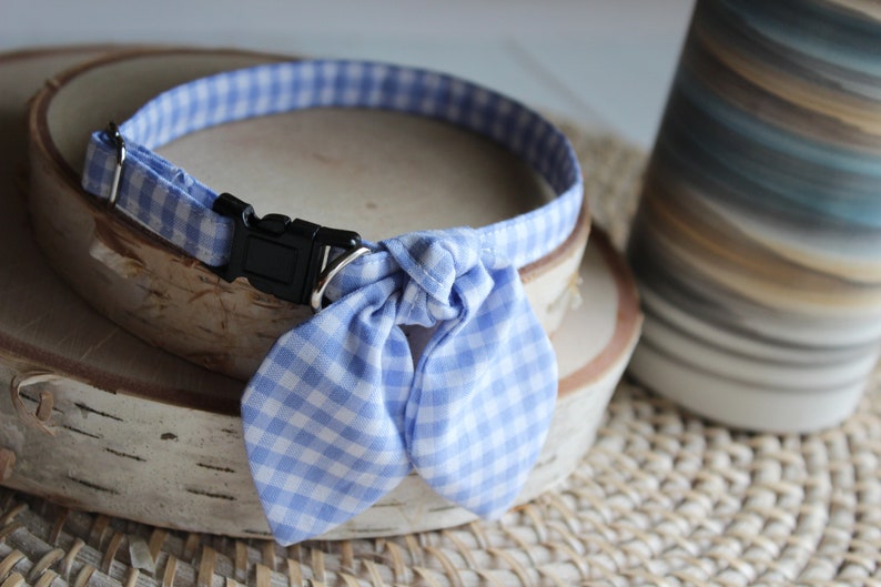 Puede incluir: Un collar de perro de gingham azul y blanco con una pajarita. El collar tiene una hebilla de pl&aacute;stico negra.