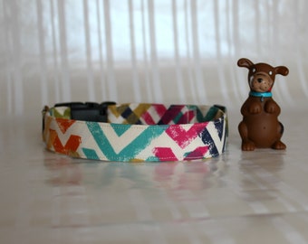 Puede incluir: Un collar de perro colorido con un patrón de chevron en tonos naranja, amarillo, turquesa, rosa y azul marino. El collar está hecho de tela y tiene una hebilla de plástico negra.