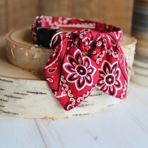 May include: Red bandana dog collar with a black buckle and a bow tie knot. The bandana has a white floral pattern.