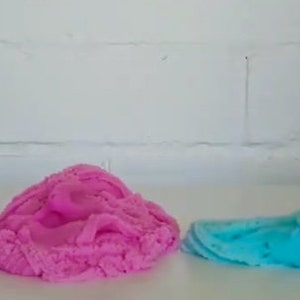 May include: Three different colors of slime, purple, pink, and blue, are shown on a white surface. The blue slime is being held by a hand.