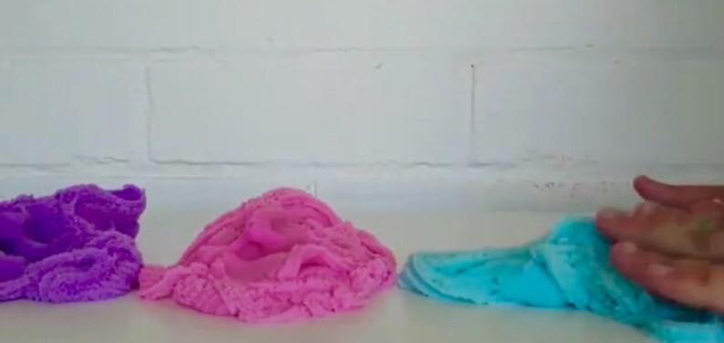 May include: Three different colors of slime, purple, pink, and blue, are shown on a white surface. The blue slime is being held by a hand.